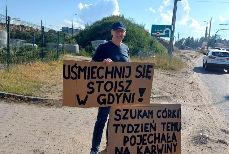 Na zdjęciu jest mężczyzna z dwoma transparentami. W rękach trzyma plakat z napisem: "Uśmiechnij się, stoisz w Gdyni", drugi plakat stoi obok i przysłania mu nogę. Napisane jest:" Szukam Córki! Tydzień temu pojechała na Karwiny".